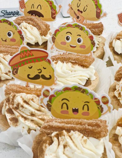 Cupcakes topped with churros and decorated with taco-themed toppers featuring various facial expressions. Some of the taco toppers are happy and smiling, one is wearing a sombrero with a mustache, and others have different expressions, creating a festive mood.